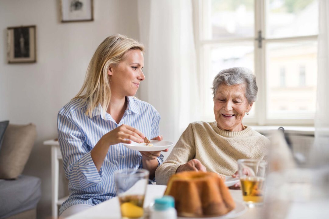 Junge Pflegerin isst gemeinsam mit älterer, pflegebedürftiger Dame
